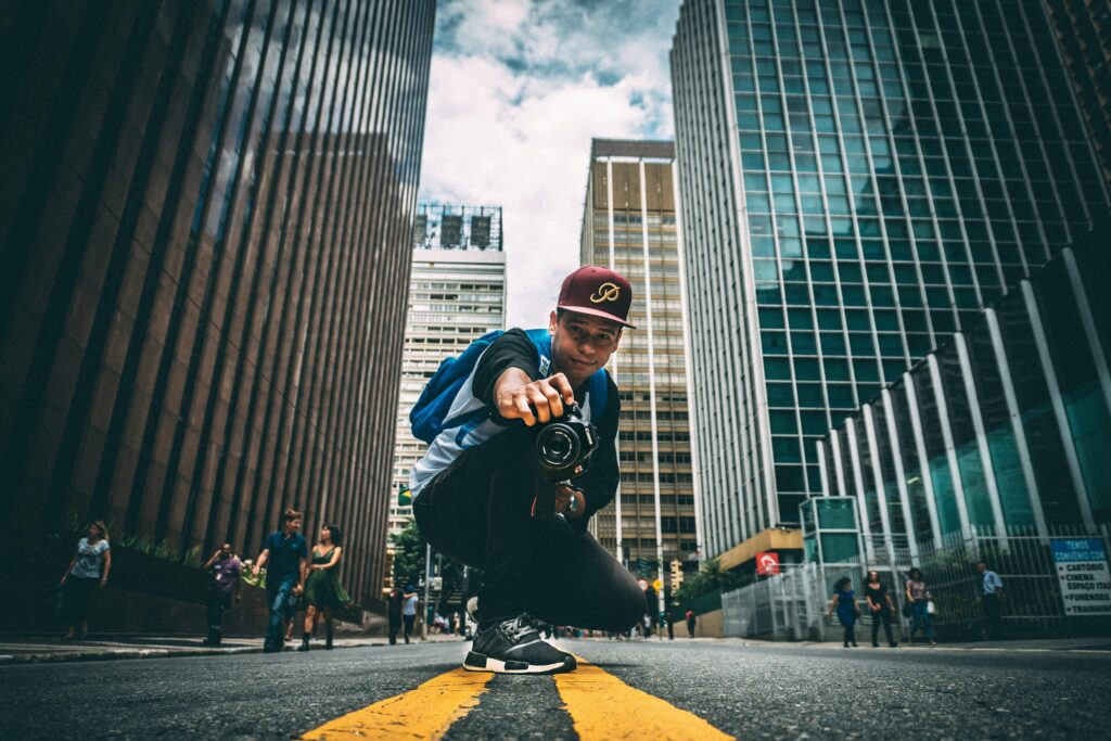 A young man with a camera kneels on a bustling city street, surrounded by skyscrapers.